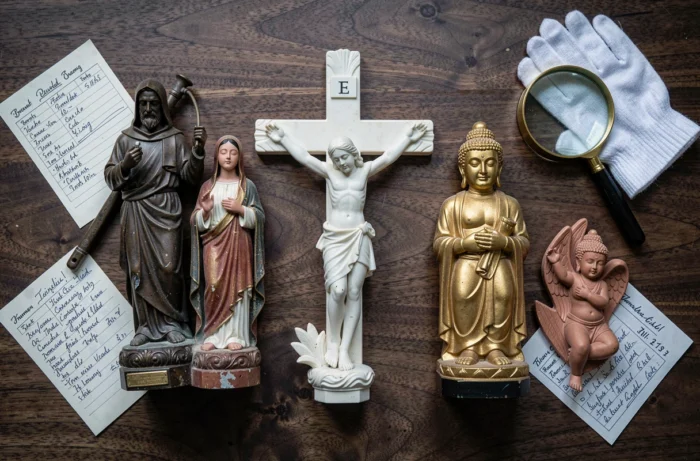 Collezione di statuette religiose antiche disposte in flat lay su un tavolo in legno scuro con guanti e schede di inventario